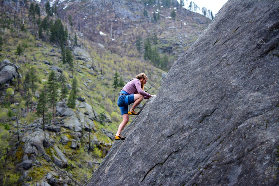 A climber in shorts skillfully ascends a steep rock face in a scenic mountainous environment.
